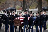 Army soldiers in ceremonial dress uniform are carrying a U.S. flag-draped coffin down a street during a funeral at a cemetery. Other soldiers are standing nearby and saluting.