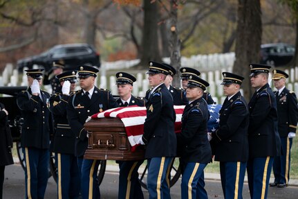 Army soldiers in ceremonial dress uniform are carrying a U.S. flag-draped coffin down a street during a funeral at a cemetery. Other soldiers are standing nearby and saluting.