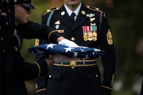 An Army Soldier wearing white gloves is holding a folded US flag in front of another Army Soldier who is wearing an Army ceremonial dress uniform.
