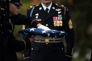 An Army Soldier wearing white gloves is holding a folded US flag in front of another Army Soldier who is wearing an Army ceremonial dress uniform.