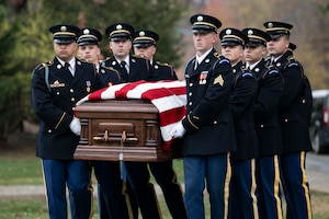 Army Soldiers in ceremonial dress uniforms are carrying a US flag-draped casket during a funeral