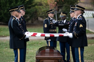 Army Soldiers wearing ceremonial dress uniforms are holding the US flag over a coffin as an Army Chaplain extends his hand over the flag while speaking during a funeral.
