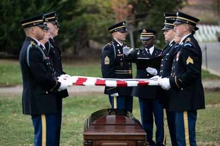 Army Soldiers wearing ceremonial dress uniforms are holding the US flag over a coffin as an Army Chaplain extends his hand over the flag while speaking during a funeral.