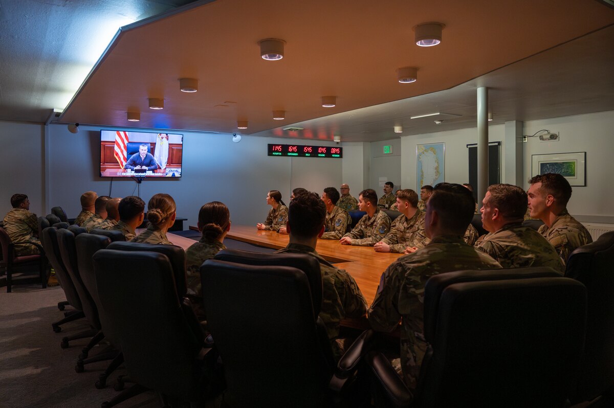 Service members sit at a conference table and look at the secretary of war on a large screen.