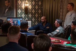 Service members gather in front of a screen to speak with the secretary of war.