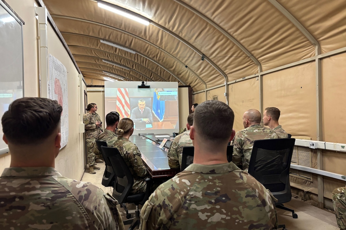 Service members gather in front of a screen to speak with the secretary of war.