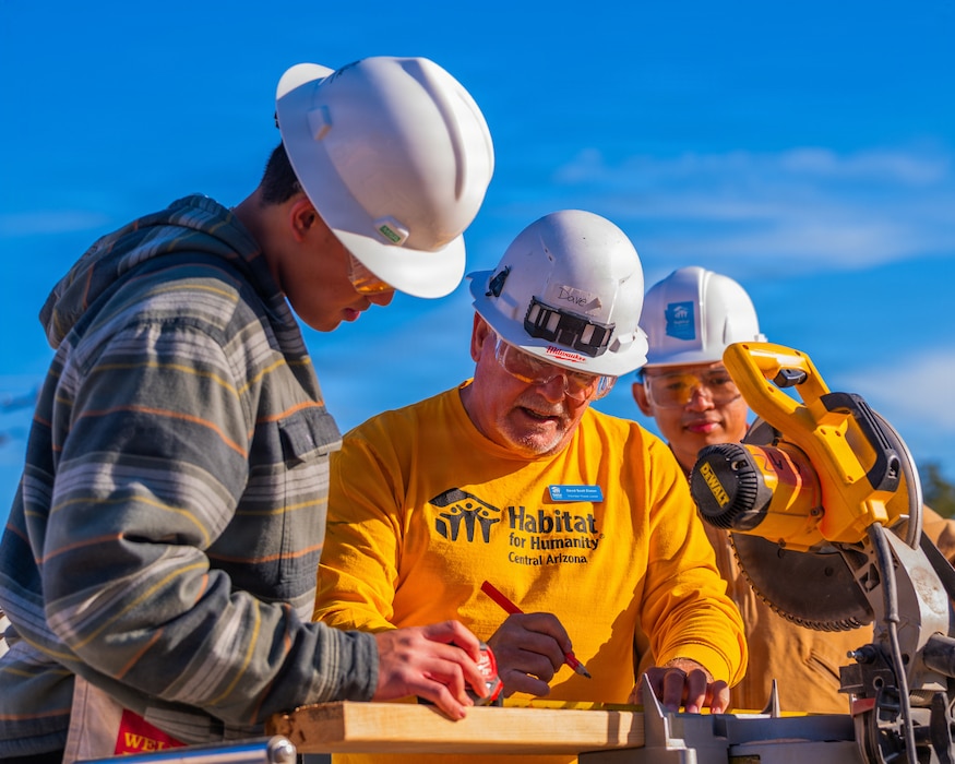 U.S. Air Force Airman 1st Class Louie Magcalas (left), 607th Air Control Squadron cyber systems technician, and Airman 1st Class Andre Castro (right), 607th Air Control Squadron cyber systems technician, are instructed by David Elston (center), Habitat for Humanity of Central Arizona volunteer house leader, on proper use of a miter saw during a Habitat for Humanity volunteer event, Dec. 13, 2025, at Buckeye, Arizona.