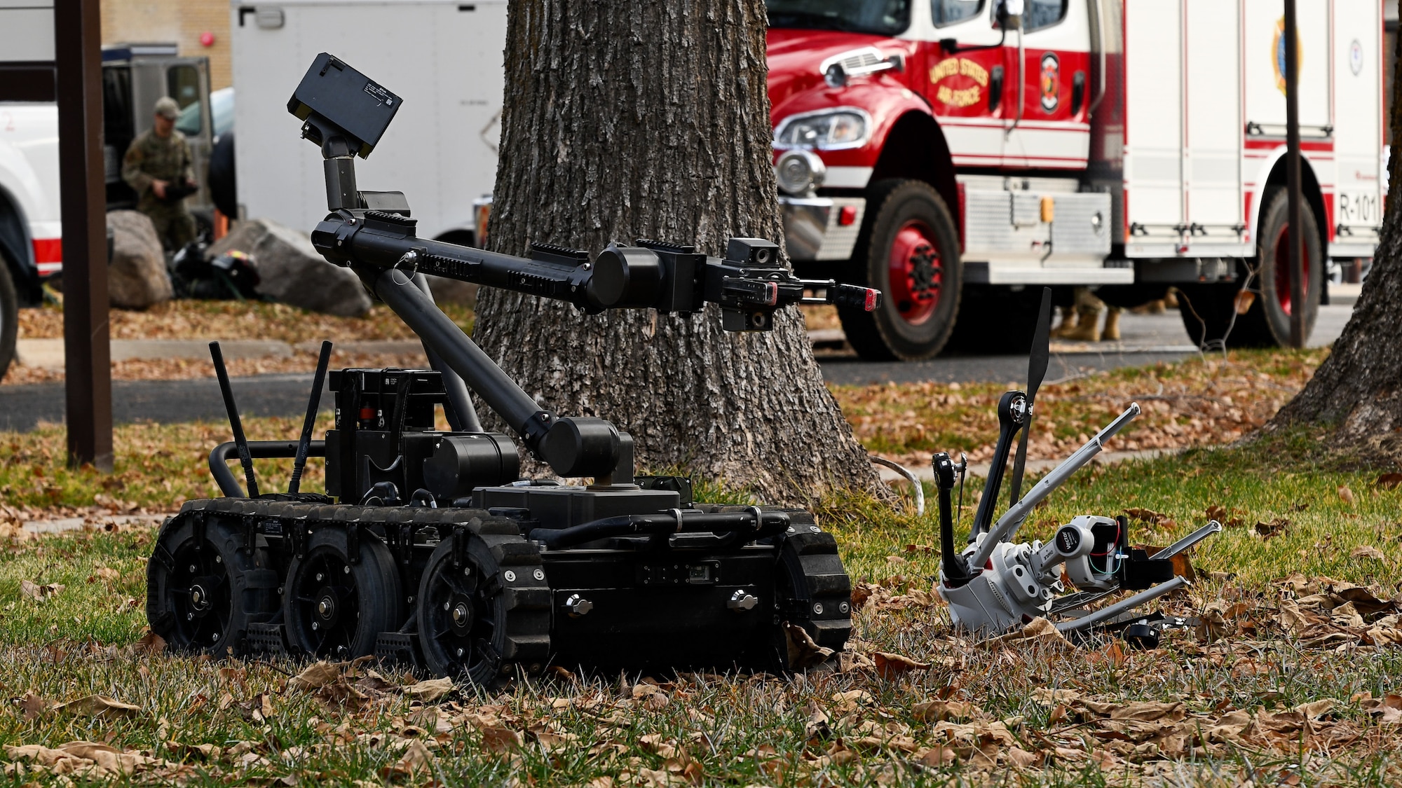 A military member in the far background next to a fire truck controls a wheeled robot that has a camera fixed to it to observe a UAS on the grass.