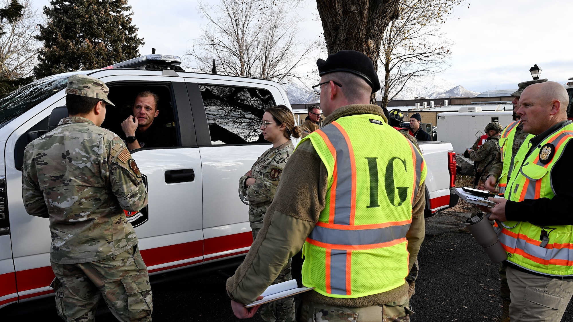 A member of Fire and Emergency Services sits in a pickup truck talking to two Airmen through his window while members donning yellow IG vests watch in the foreground.