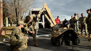 Two Airmen study the protruding arm of a wheeled robot while other exercise players watch in the background.