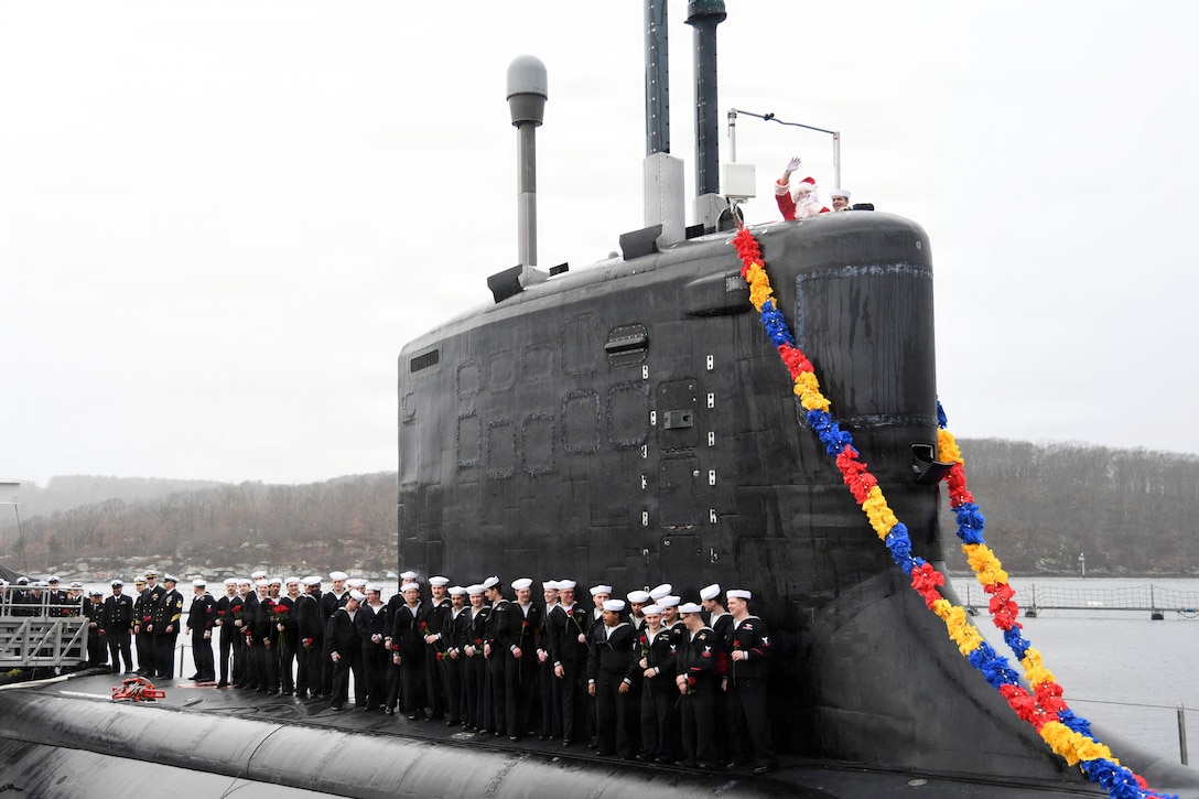 Sailors in ceremonial dress stand in formation aboard a submarine as Santa waves to the right on a foggy day.