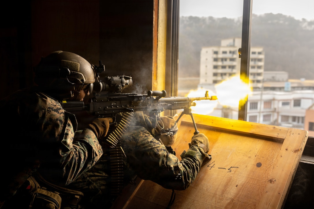 Two Marines lean on a wooden surface to fire weapons outside of a window, creating fireballs.