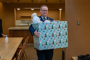 An Airman wearing their dress uniform smiles while carrying a box of toys.