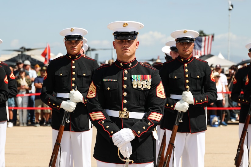 Marines wearing formal black attire and white caps stand at attention with weapons during a synchronized march outdoors during the day.