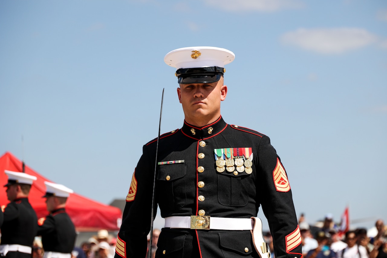 A Marine wearing formal black attire and a white cap stands at attention outside during the day.