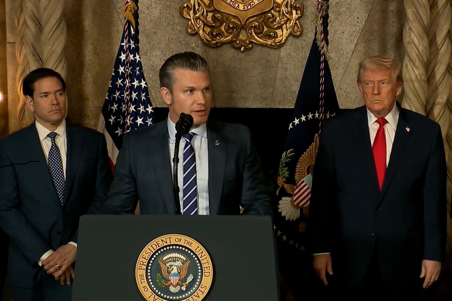 A man in business attire stands and speaks at a lectern with the American flag to the left behind him. Three men in similar attire stand behind him.