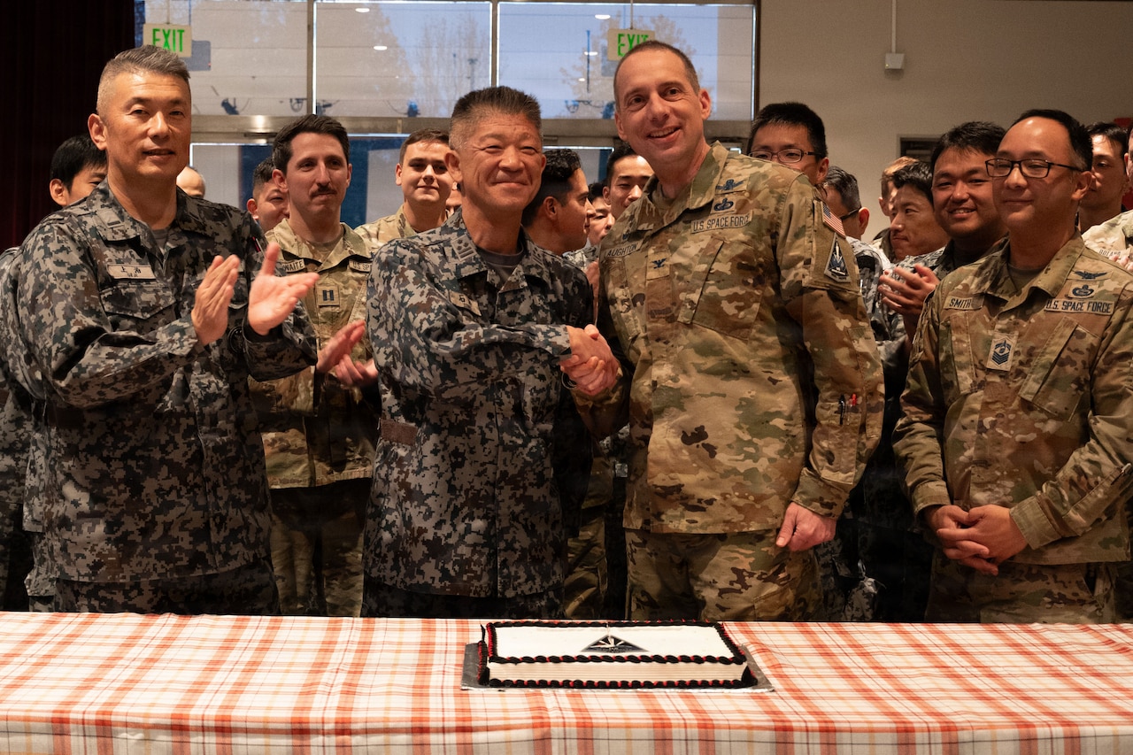 Two men in military camouflage uniforms shake hands while standing behind a table with a cake on it. They are surrounded by other service members in similar attire, who are clapping.
