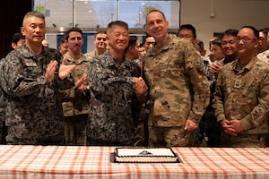 Two men in military camouflage uniforms shake hands while standing behind a table with a cake on it. They are surrounded by other service members in similar attire, who are clapping.