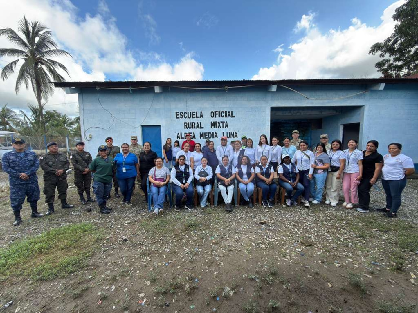 People in military uniforms and civilian clothing gather for a photo in front of a school.