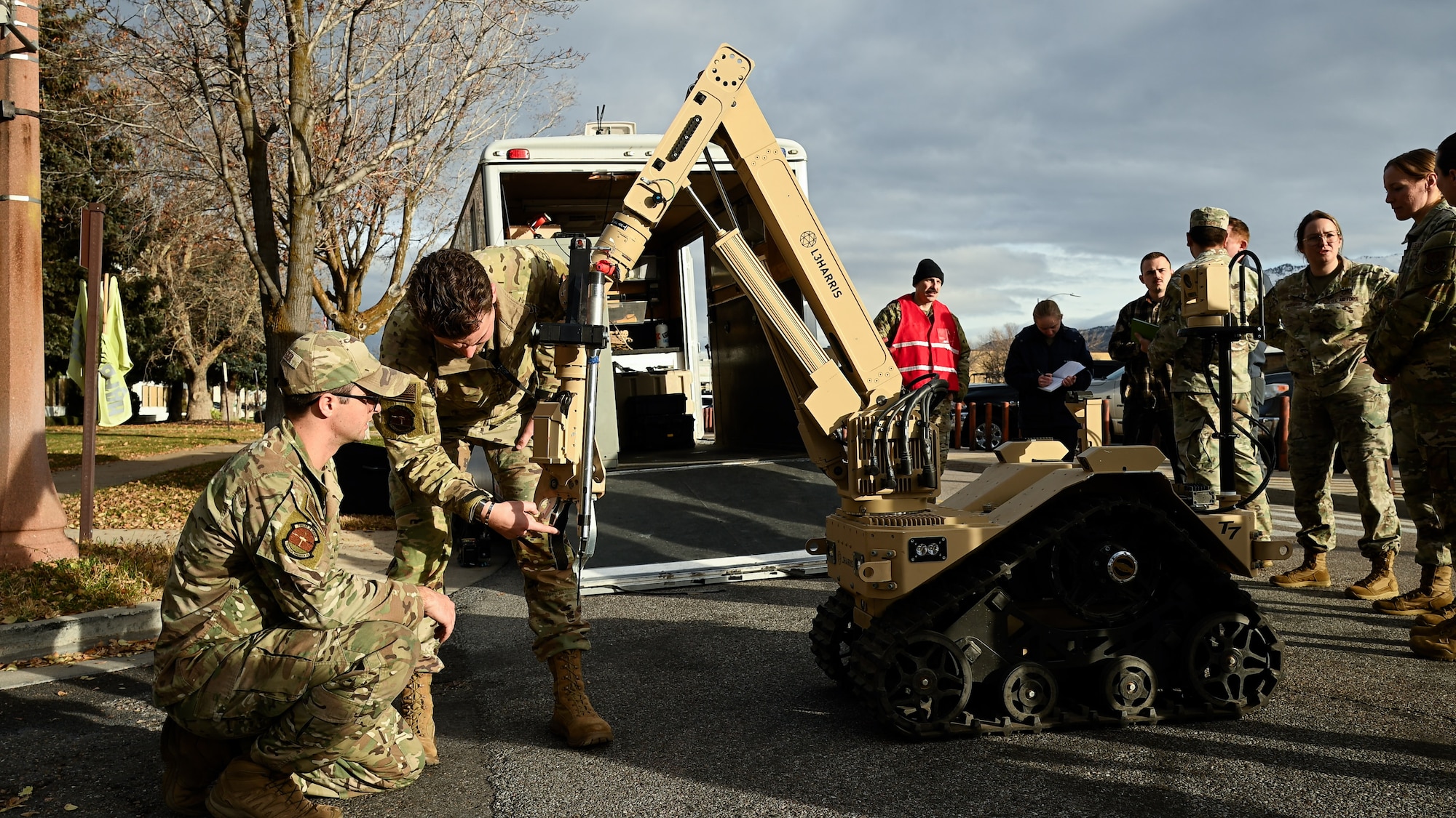 Two Airmen study the protruding arm of a wheeled robot while other exercise players watch in the background.