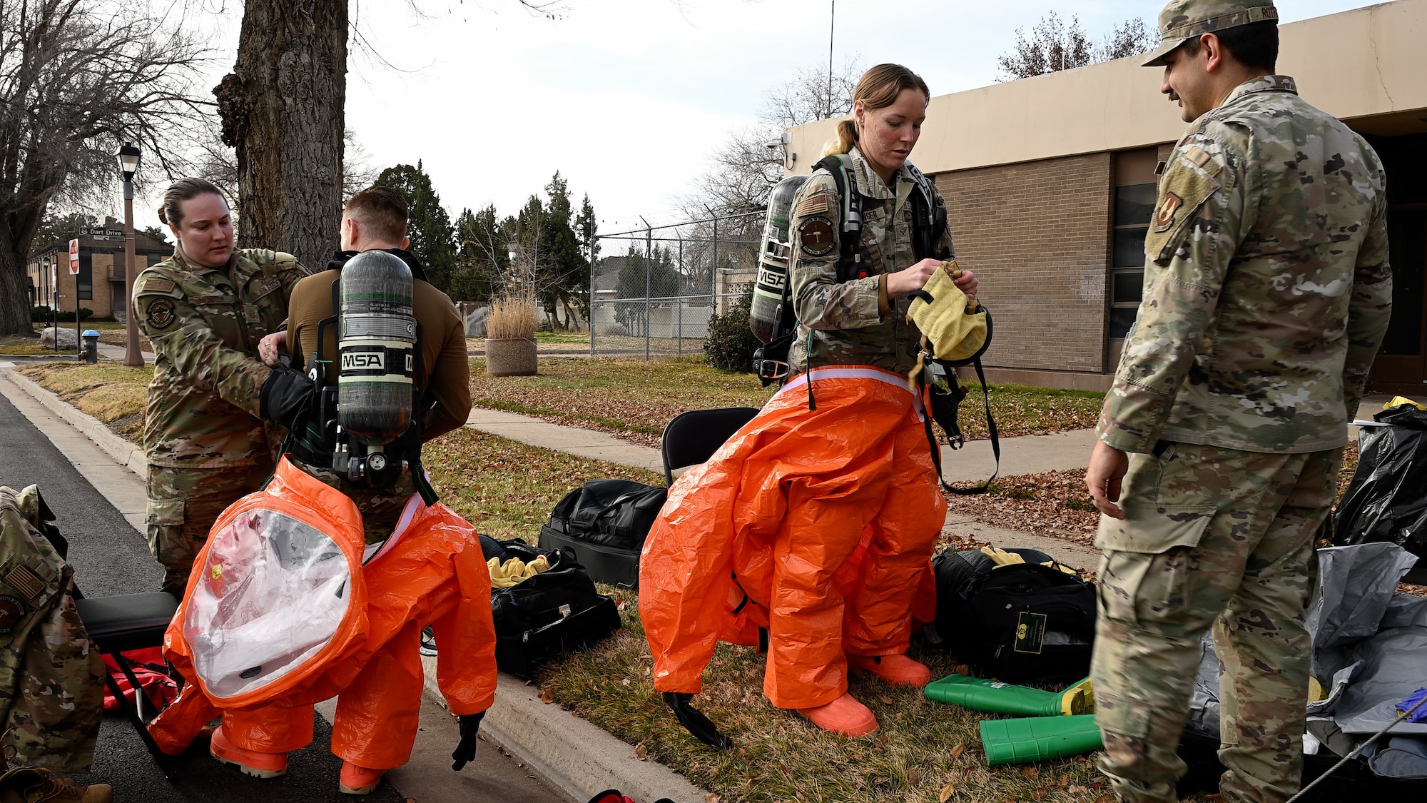 Two military members suit up in orange protective suits with the assistance of two other military members.