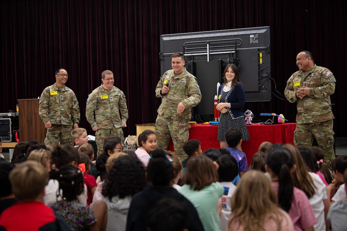 Four service members in military camouflage uniforms and a woman in business attire smile while standing in front of a group of seated children.
