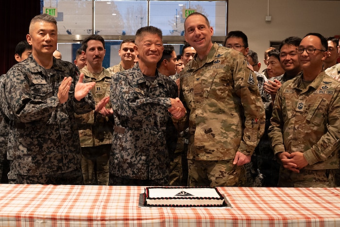 Two men in military camouflage uniforms shake hands while standing behind a table with a cake on it. They are surrounded by other service members in similar attire, who are clapping.