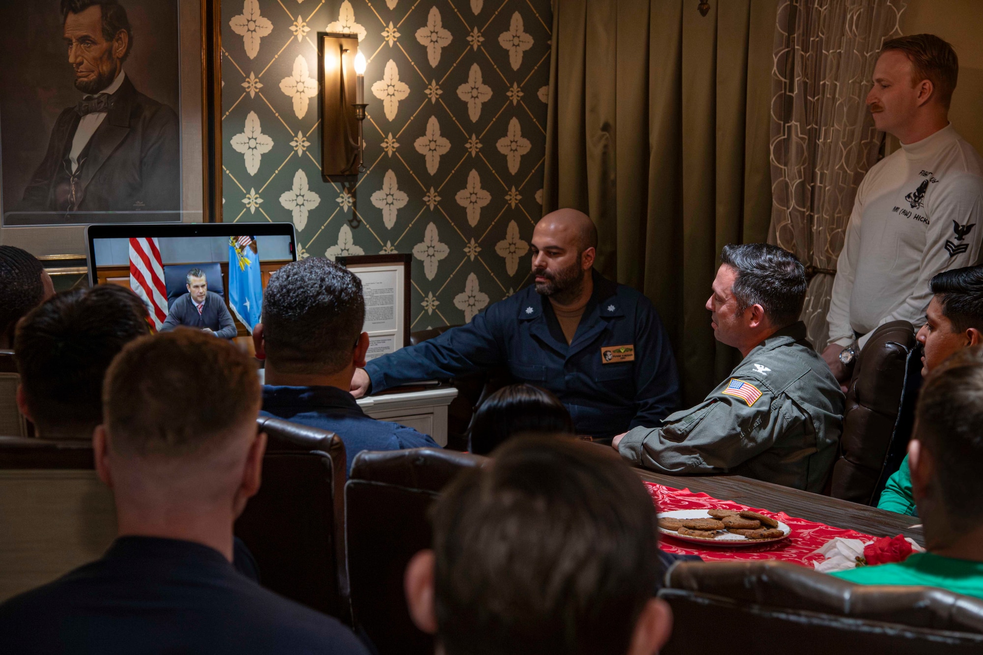 Service members gather in front of a screen to speak with the secretary of war.