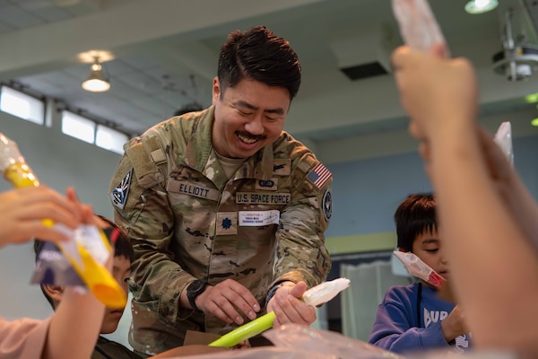 A man in a military camouflage uniform smiles while holding a model rocket. He is standing at a table with children as they put together model rockets.