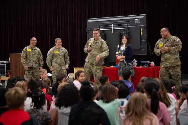 Four service members in military camouflage uniforms and a woman in business attire smile while standing in front of a group of seated children.