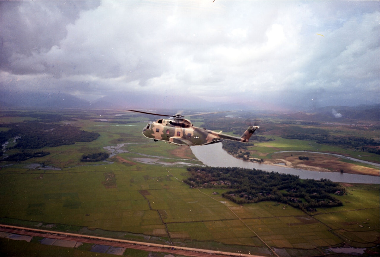 A military helicopter flies over green flatlands and a river.