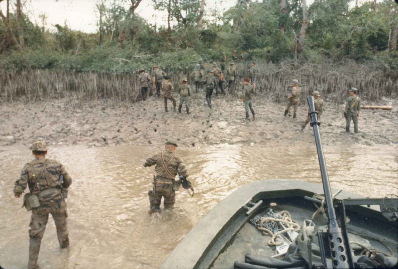 Two men in military camouflage uniforms wade through shallow water to get to a shore from a small boat. Others in similar attire are on the beachhead.
