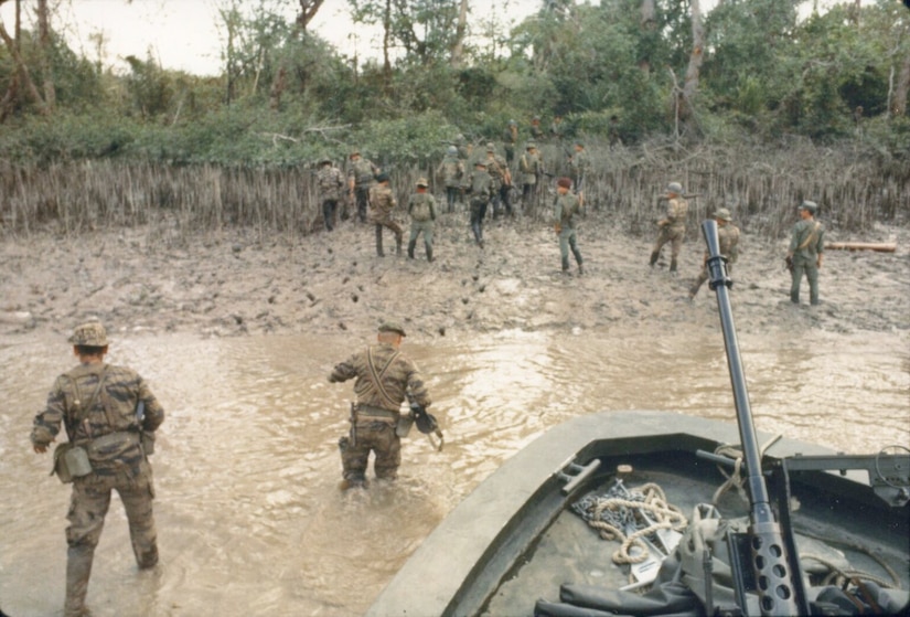 Two men in military camouflage uniforms wade through shallow water to get to a shore from a small boat. Others in similar attire are on the beachhead.