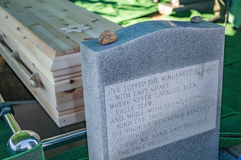 A headstone is shown with an engraving on it and a casket behind it.