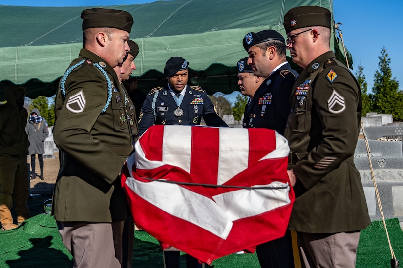 Seven service members in military dress uniforms lower a casket draped with the American flag into a grave.