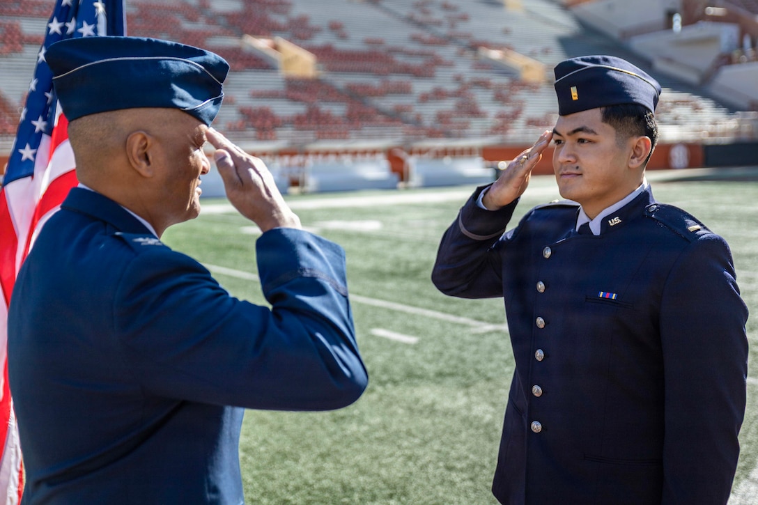 A father and son in ceremonial dress face each other while saluting on a stadium field during the day.