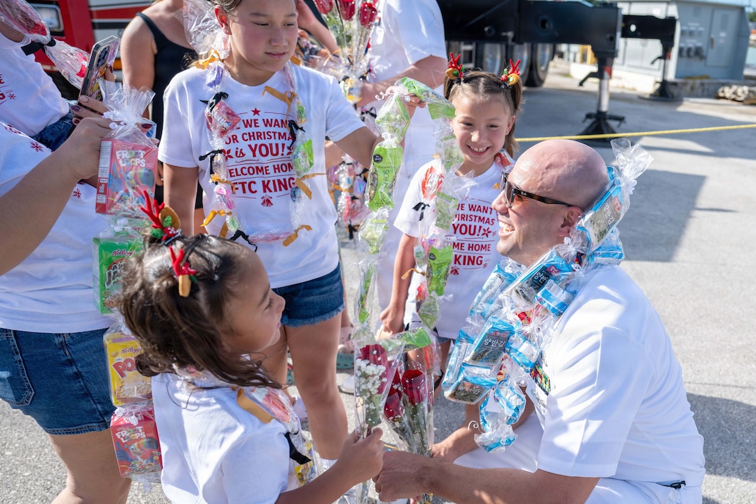 A kneeling sailor wearing a necklace made of snacks smiles while greeting three children dressed in matching T-shirts.