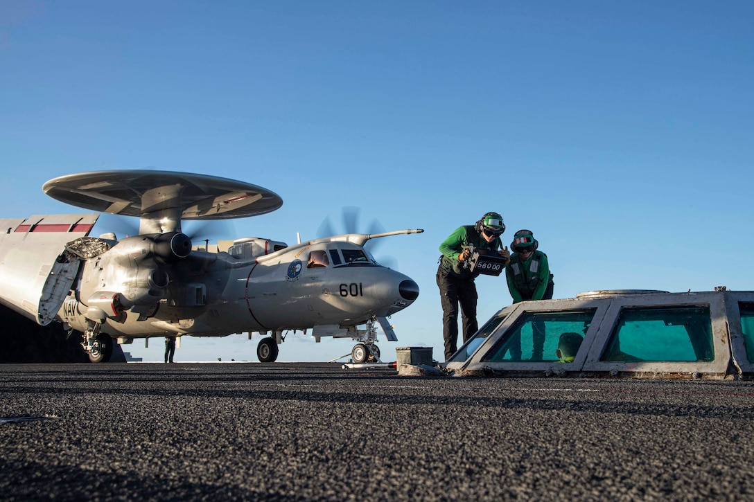 Two sailors work on an aircraft on a ship as another aircraft sits in the background against a blue sky.