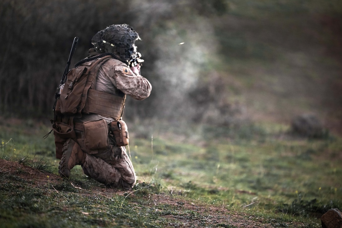 A Marine in tactical gear kneels while firing a weapon in a wooded area.