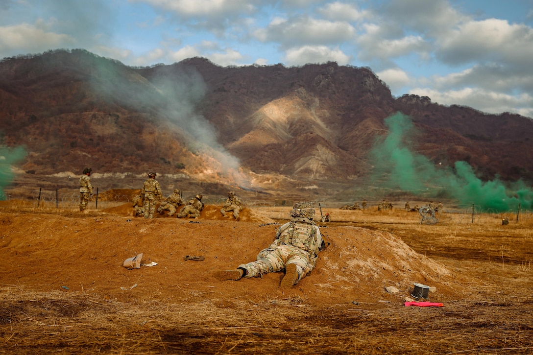 Clouds of green smoke hover over soldiers in tactical gear as they lie in the dirt near a mountain during the day.