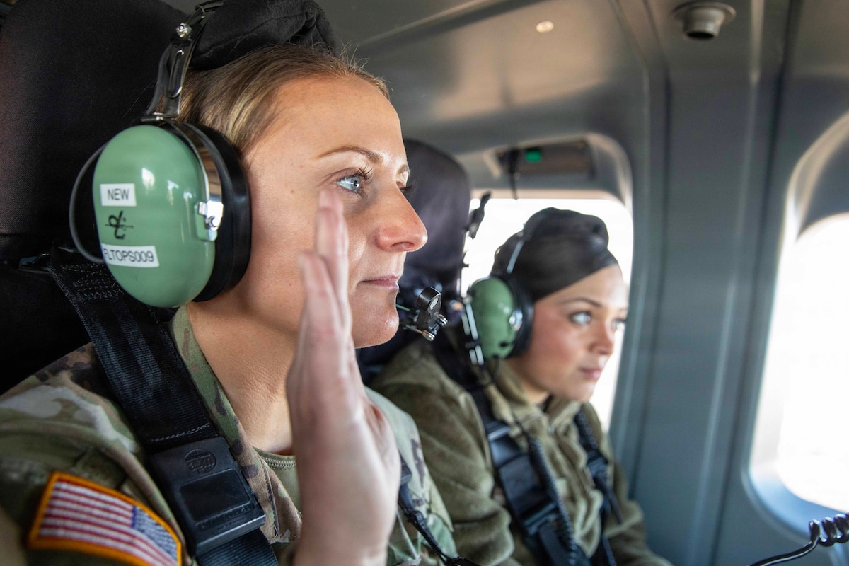 A soldier wearing a headset raises their right hand while sitting next to a fellow service member in a helicopter.