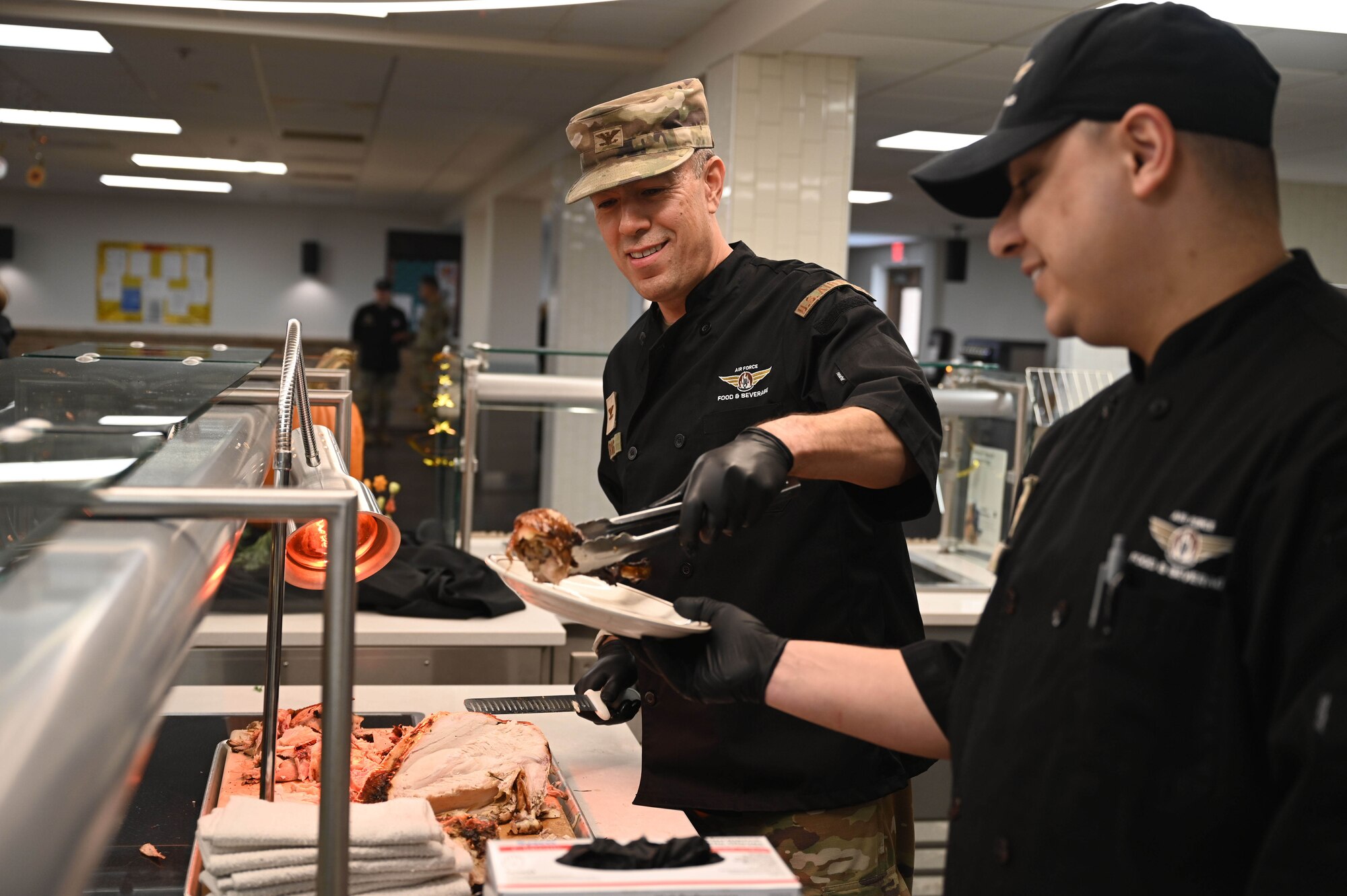 U.S. Air Force Col. Daniel Craig, 355th Mission Support Group Commander, left, and Senior Airman Brandon Cortez, 355th Force Support Squadron food services journeyman, carve and serve turkey during a Thanksgiving day meal in the Desert Inn Dining Facility at Davis-Monthan Air Force Base, Arizona, Nov. 27, 2025. Senior leaders serving personnel a fresh cooked holiday meal is a long-standing military tradition for U.S. dining facilities across the Air Force on Thanksgiving Day and Christmas Day. (U.S. Air Force photo by Staff Sgt. Kahdija Slaughter)