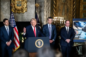 A man in a suit with a red tie talks from a lectern indoors, as three other men stand behind him.