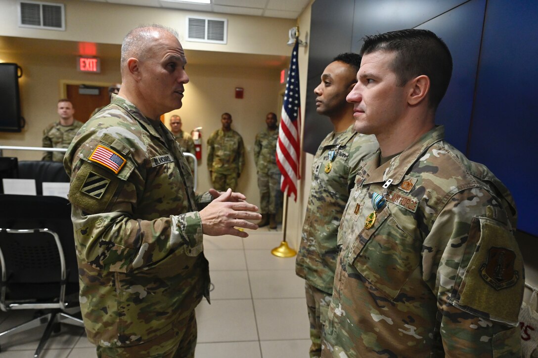 A solider speaks to an airman and a soldier in a room with a blue wall and a standing American flag.