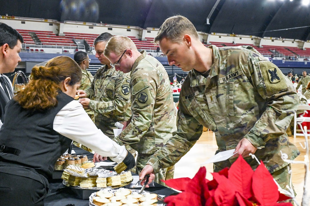 Four guardsmen fill their plates at a dessert table as two people help with red and blue bleachers shown in the distance behind.