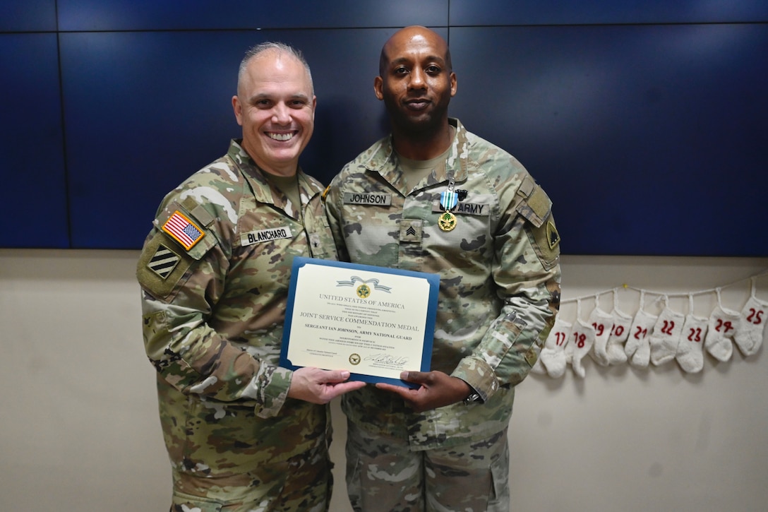 Two soldiers hold up an award and pose for a picture in front of a dark blue and white wall.