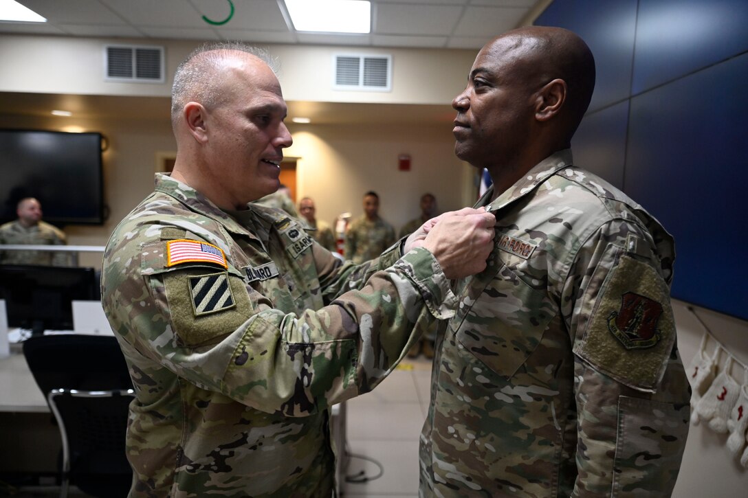 A soldier pins an award on the lapel of an airman in a room with other service members standing in the distance.