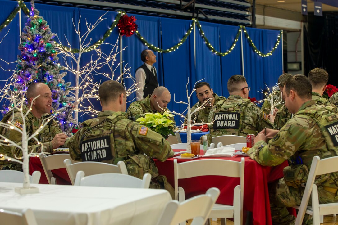 Guardsmen eat at tables covered with red tablecloth in front of lights, a Christmas tree and other decorations in front of a blue curtained divider.