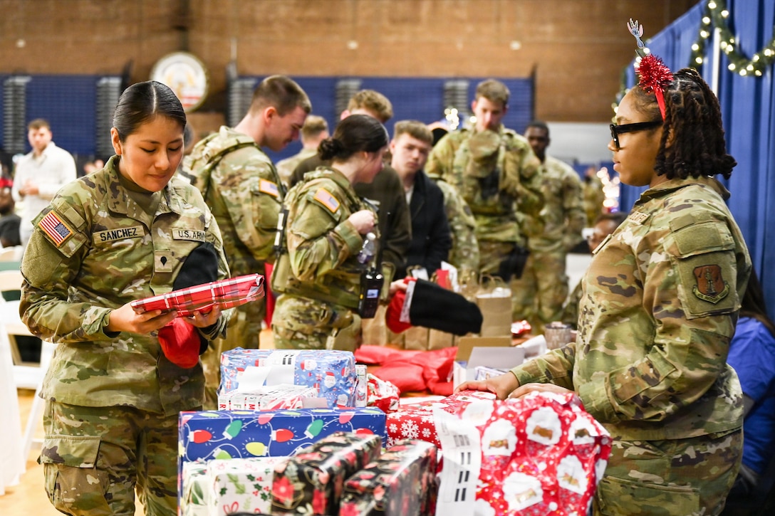 Two soldiers stand in front of a table filled with presents while other soldiers stand behind.