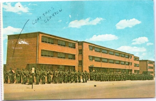 A three-story red brick building with soldiers in green uniforms lined up in front and a blue sky in the background.